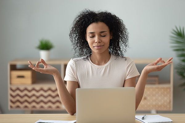 African woman meditating sit at desk in front of pc, serene mixed-race female closed eyes folded fingers mudra symbol do exercise practising yoga reducing anxiety stress positive frame of mind concept